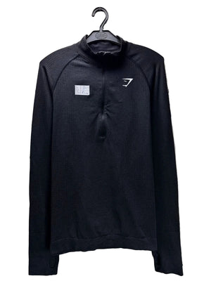 A black long-sleeve quarter-zip athletic pullover with a textured waffle knit pattern, hanging on a black hanger against a background of printed newspaper pages. The left chest features a small white rectangular patch, and the right chest features a white stylized logo.