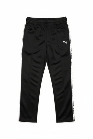 Black Puma track pants hanging on a black hanger against a background of newspaper pages. The pants feature a wide elastic waistband, a small white Puma logo on the upper left thigh, and a prominent white Puma stripe running down the side of the leg.