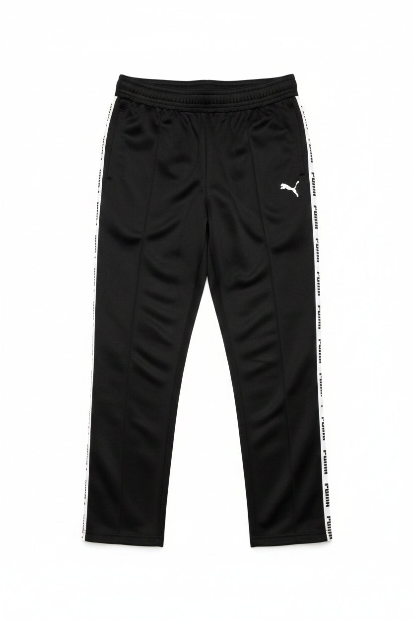 Black Puma track pants hanging on a black hanger against a background of newspaper pages. The pants feature a wide elastic waistband, a small white Puma logo on the upper left thigh, and a prominent white Puma stripe running down the side of the leg.